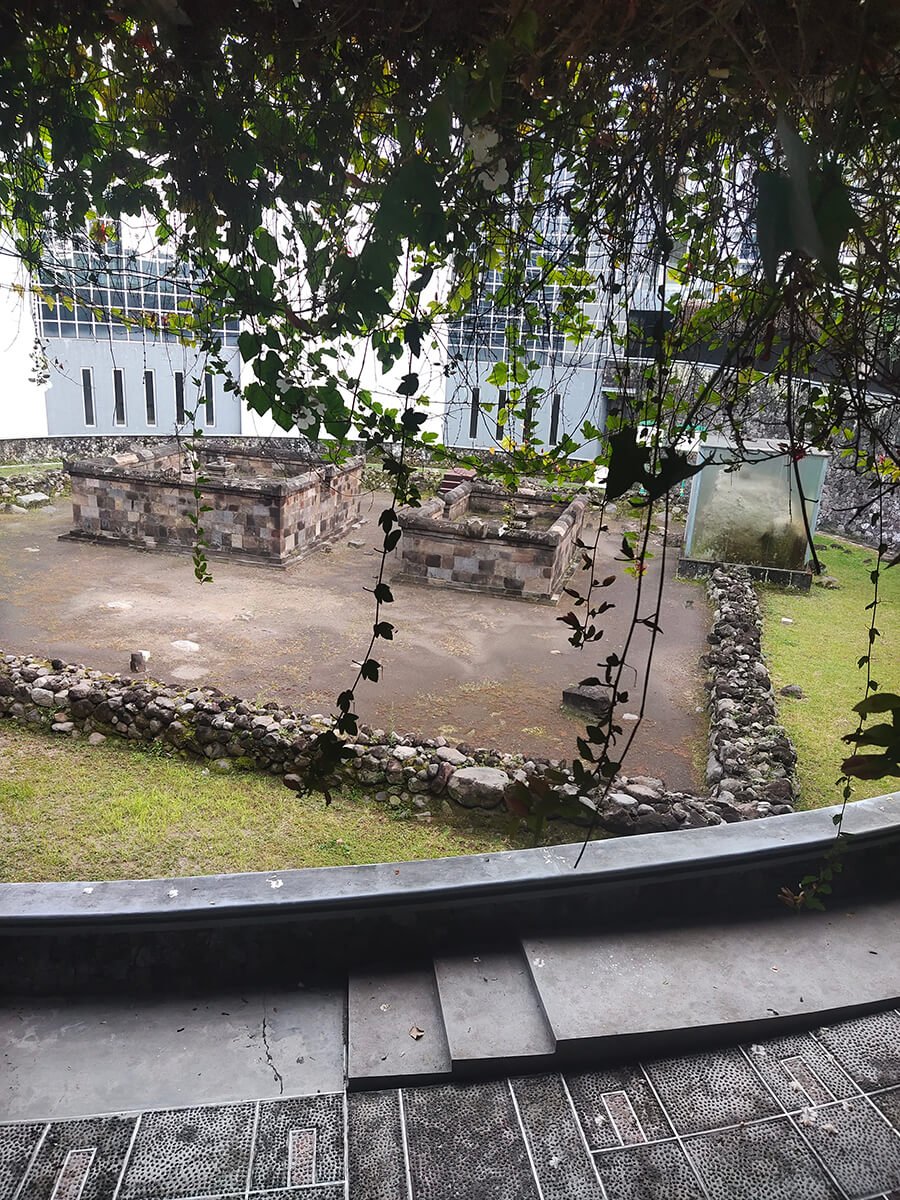 View of Kimpulan Temple ruins seen through hanging vines, located at the UII campus in Yogyakarta, featuring ancient stone structures and a glass-protected artifact.