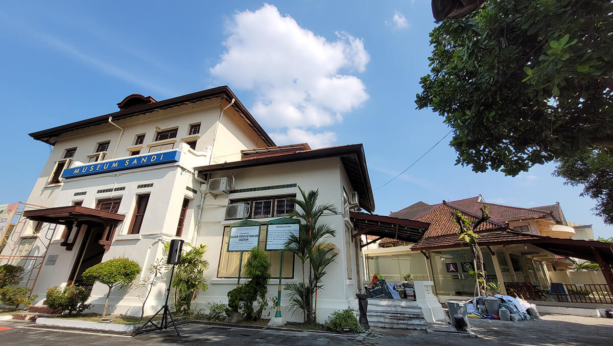 A white colonial-style building with a sign that reads "Museum Sandi" on the front, under a blue sky with a few clouds.