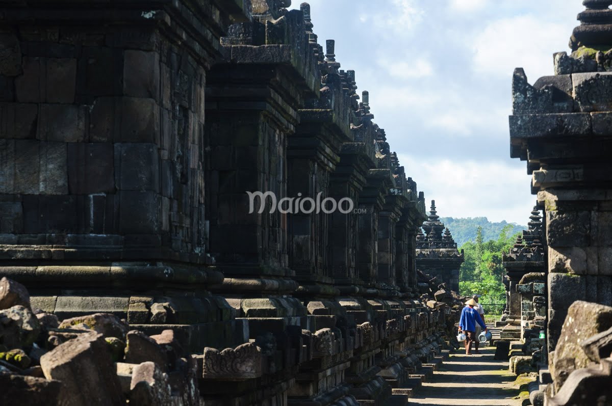 A narrow path between ancient stone structures at Plaosan Temple complex with a person walking in the distance.