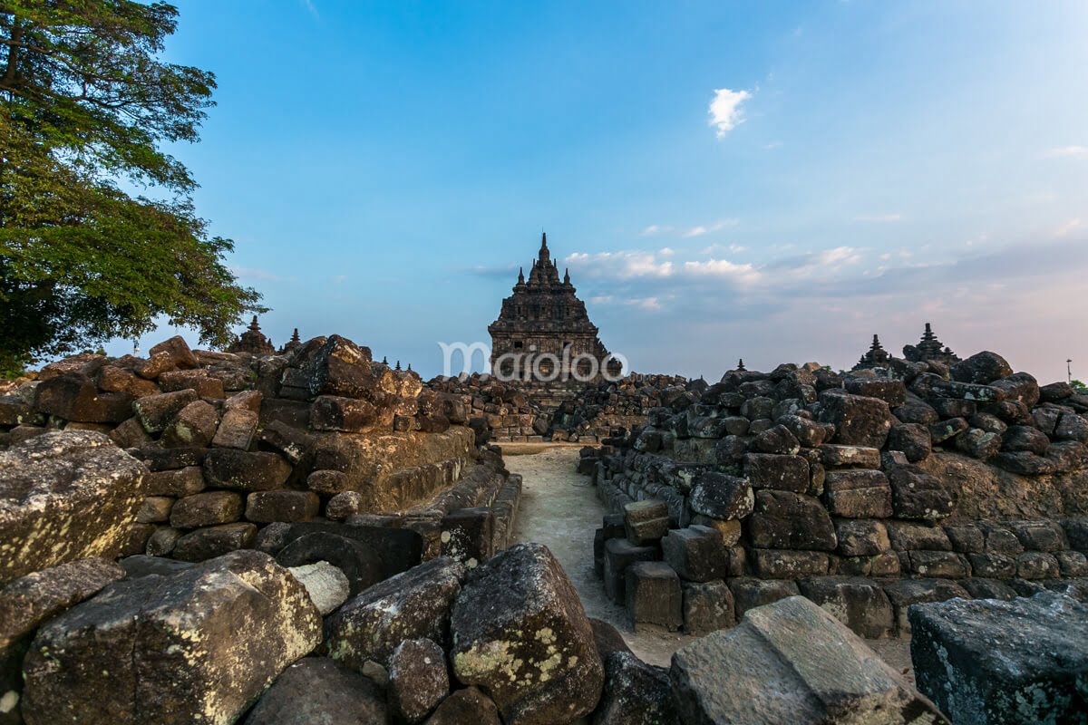 The main structure of Plaosan Temple seen through piles of ancient stones and a clear blue sky.