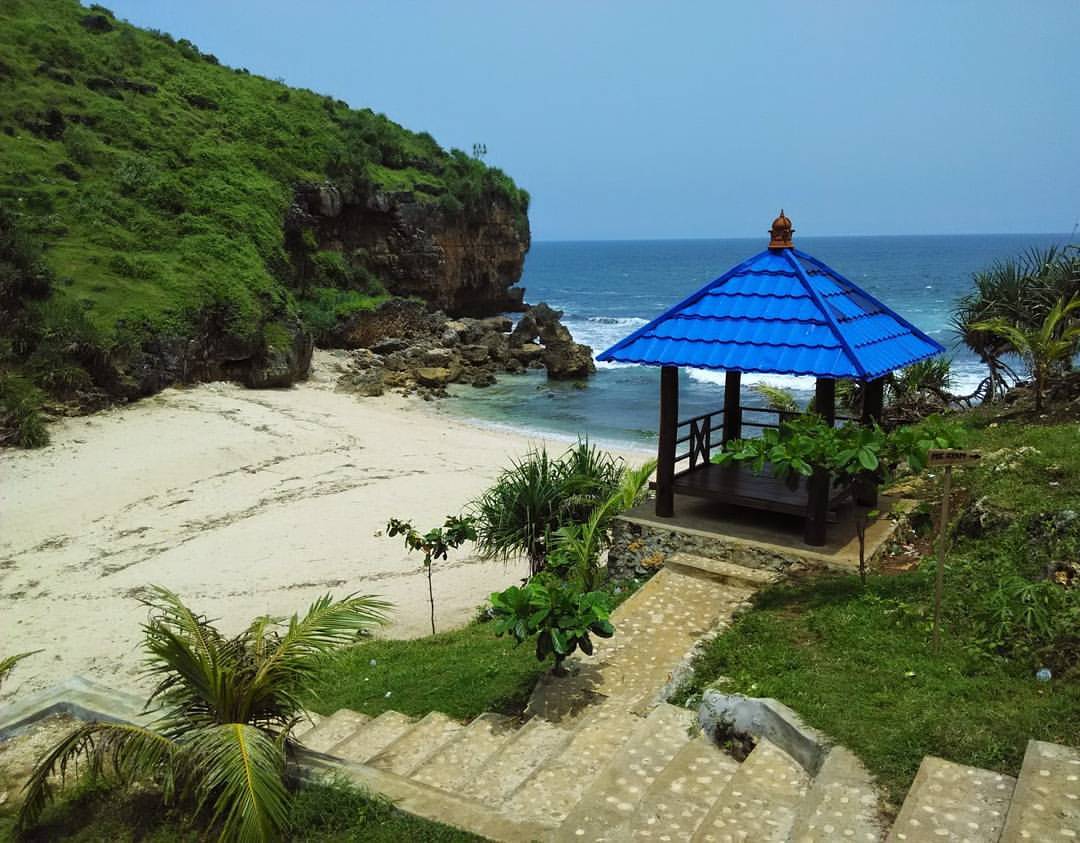 A small pavilion with a blue roof near the sandy shore of Ngedan Beach, surrounded by green hills and rocky terrain.