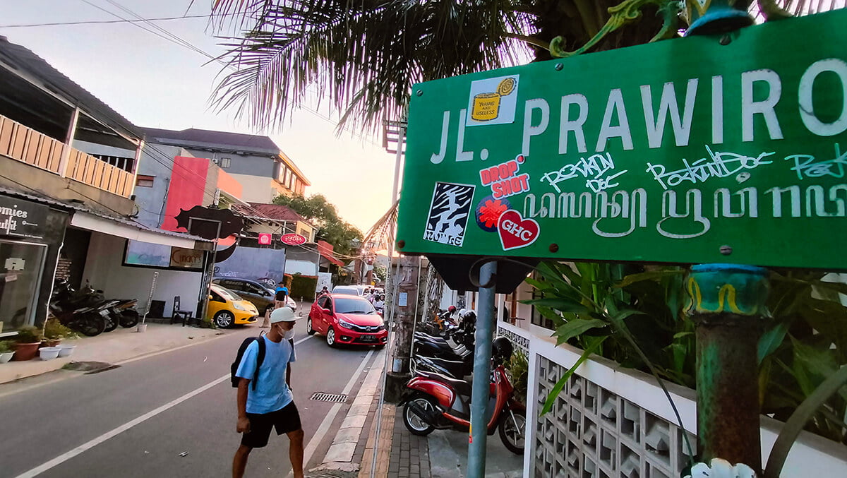 A street sign for Prawirotaman street with people and vehicles in the background.