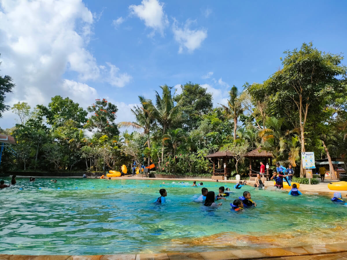 People swimming and playing in a pool surrounded by trees at Waterboom Jogja.