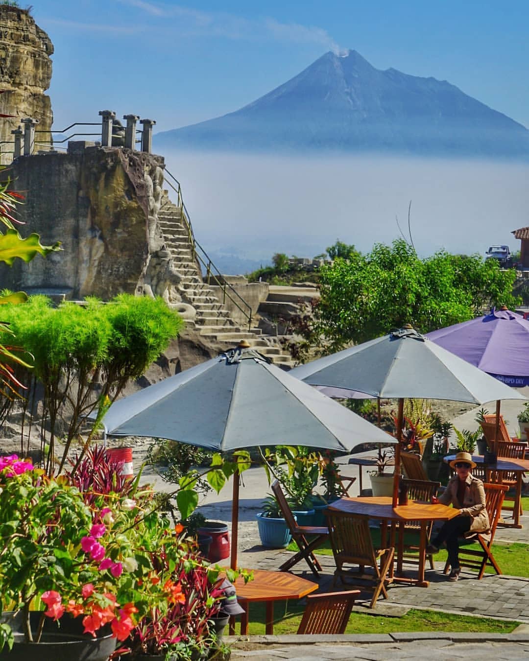 Visitor sitting at a table under an umbrella with Breksi Cliff and Mount Merapi in the background.