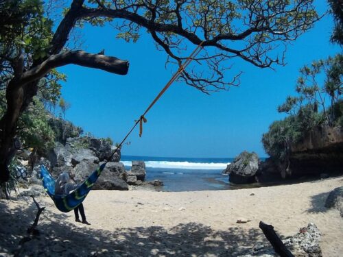 A person relaxing on a hammock under a tree at Lolang Beach with clear blue skies and rocky cliffs in the background.