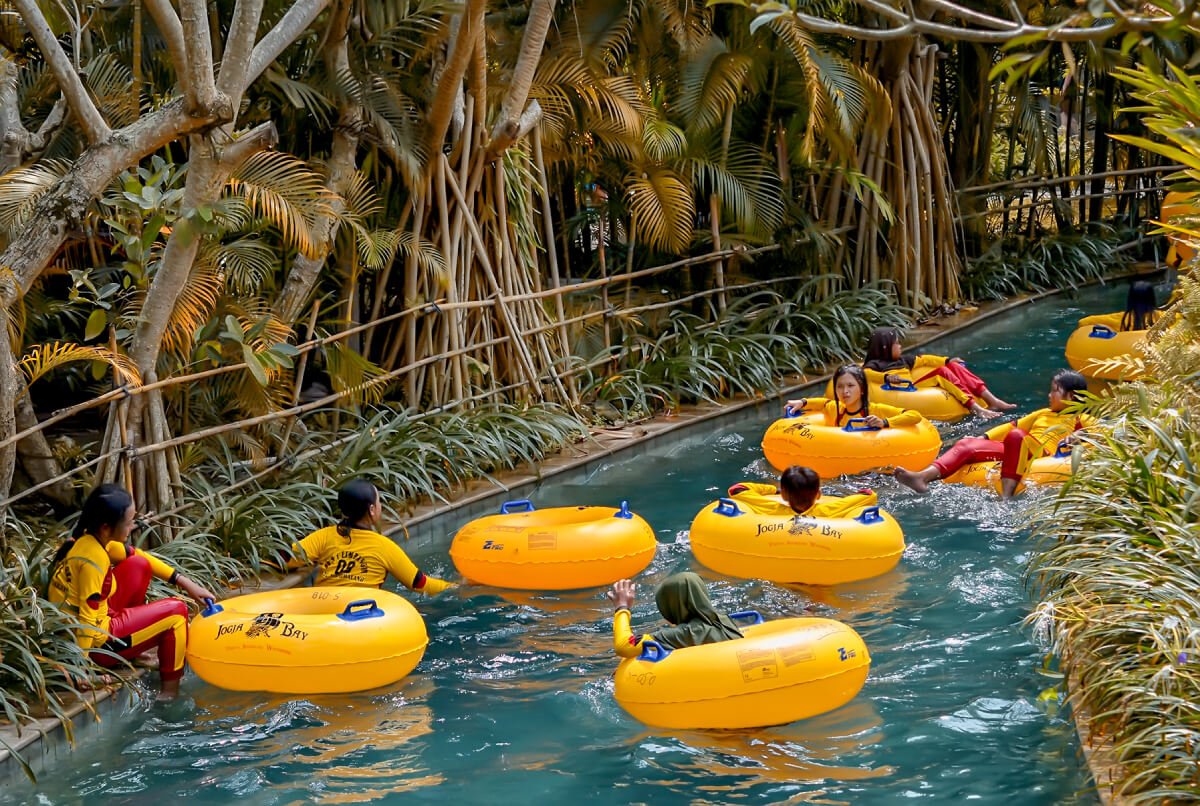 People floating on yellow inner tubes in a lazy river surrounded by trees at Waterboom Jogja.