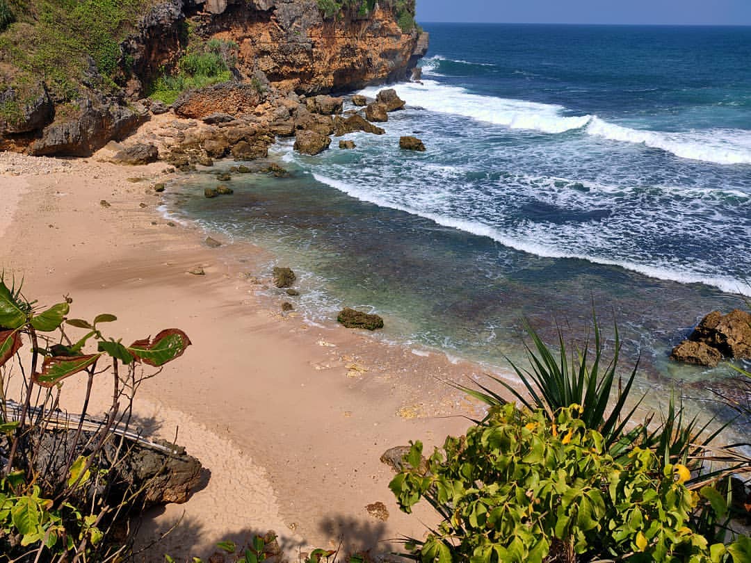 A rocky part of Ngedan Beach with clear blue waves crashing onto the sandy shore and surrounding cliffs.