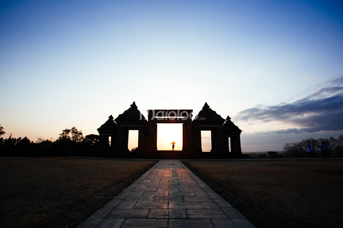 A person stands under the gate of Ratu Boko Palace with the sun setting behind them.