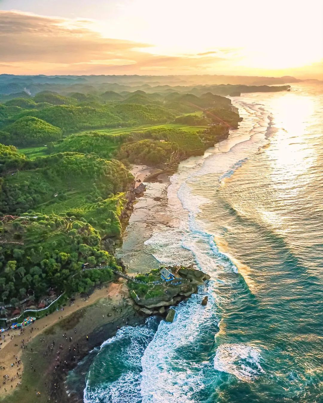 Aerial view of Kukup Beach at sunset with waves crashing on the shore and green hills in the background.