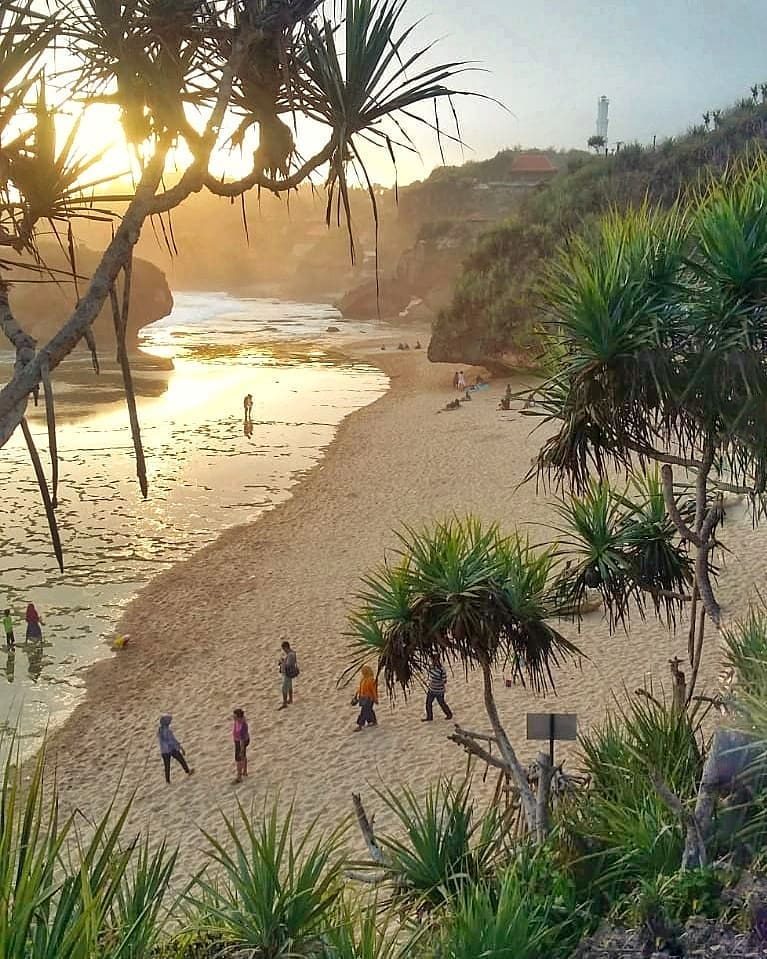 People walking on the sandy shore of Kukup Beach during sunset, framed by tropical trees.