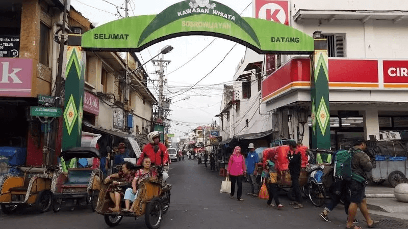 People walking and riding becaks under the green Sosrowijayan archway.