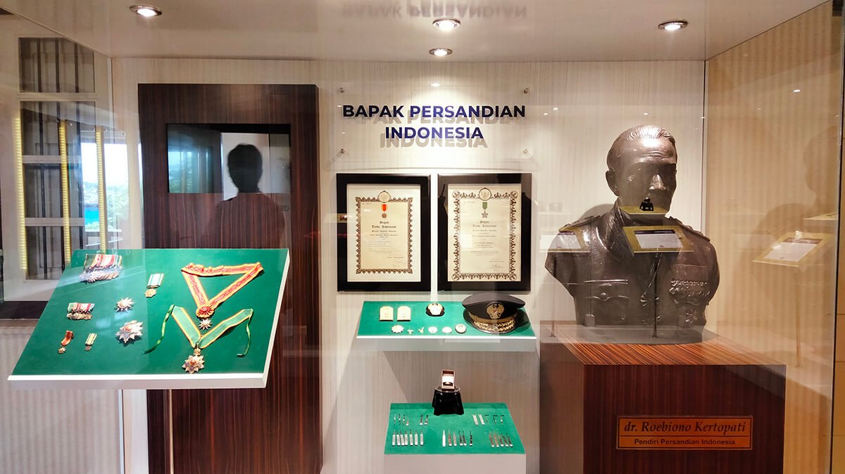 A museum display featuring medals, certificates, and a bust of dr. Roebiono Kertopati titled "Bapak Persandian Indonesia."