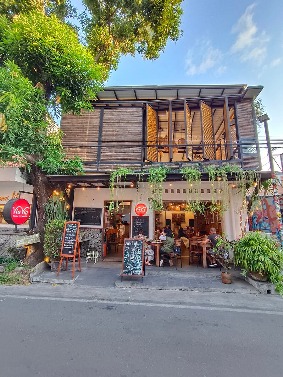 The exterior of ViaVia Cafe with customers seated inside and outside, and green plants hanging from the roof.