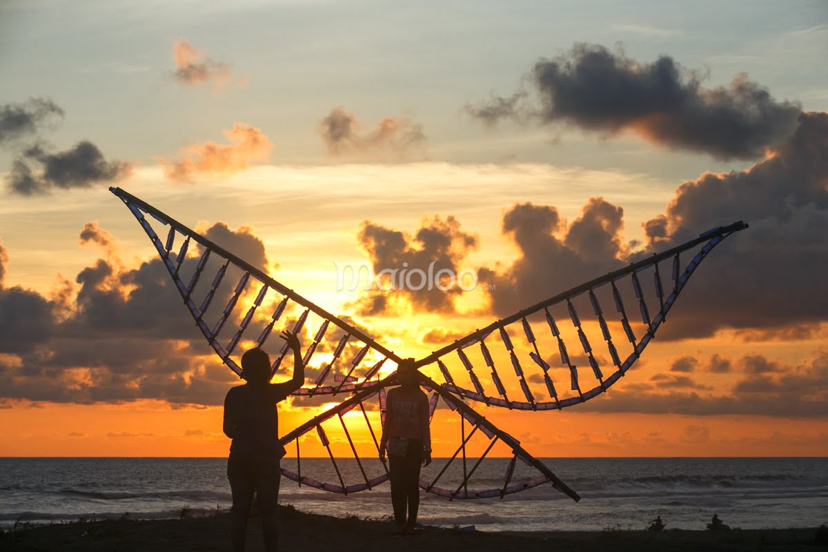 Two visitors posing by a wing-like installation made of recycled bottles at sunset.