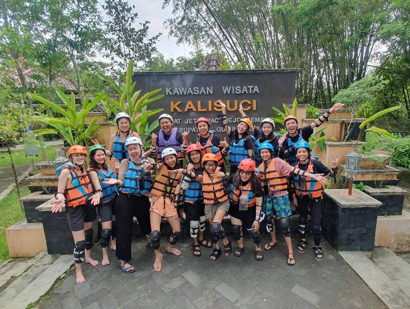 A group of people in life jackets and helmets posing happily in front of the Kalisuci Cave Tubing sign.