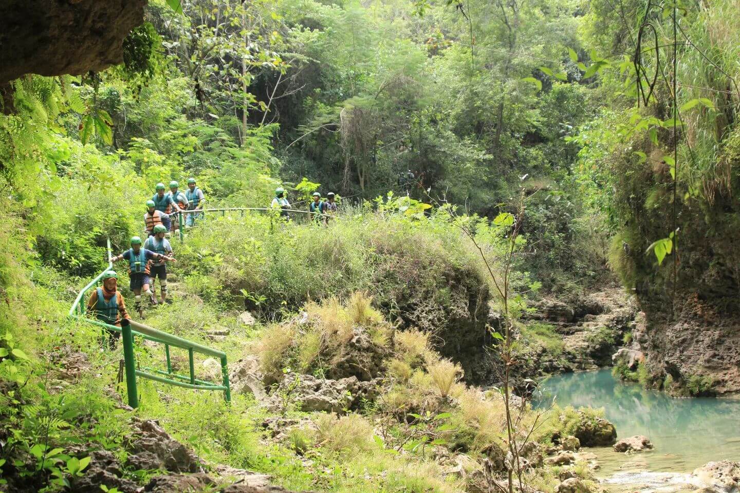 A group of people walking down a green pathway leading to the Kalisuci River and caves, surrounded by lush vegetation.