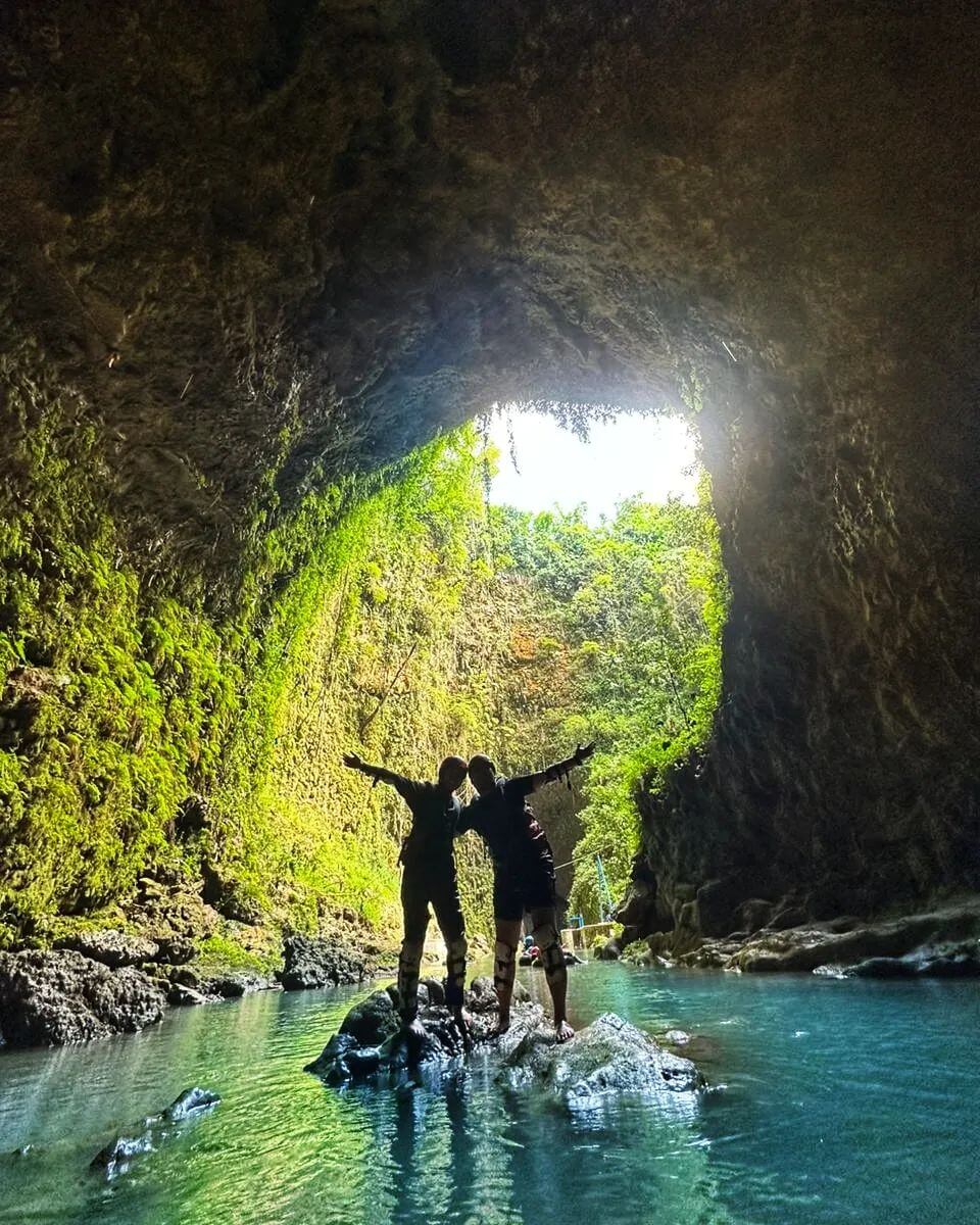 Two people standing on rocks in a sunlit cave at Kalisuci, surrounded by greenery and clear water.