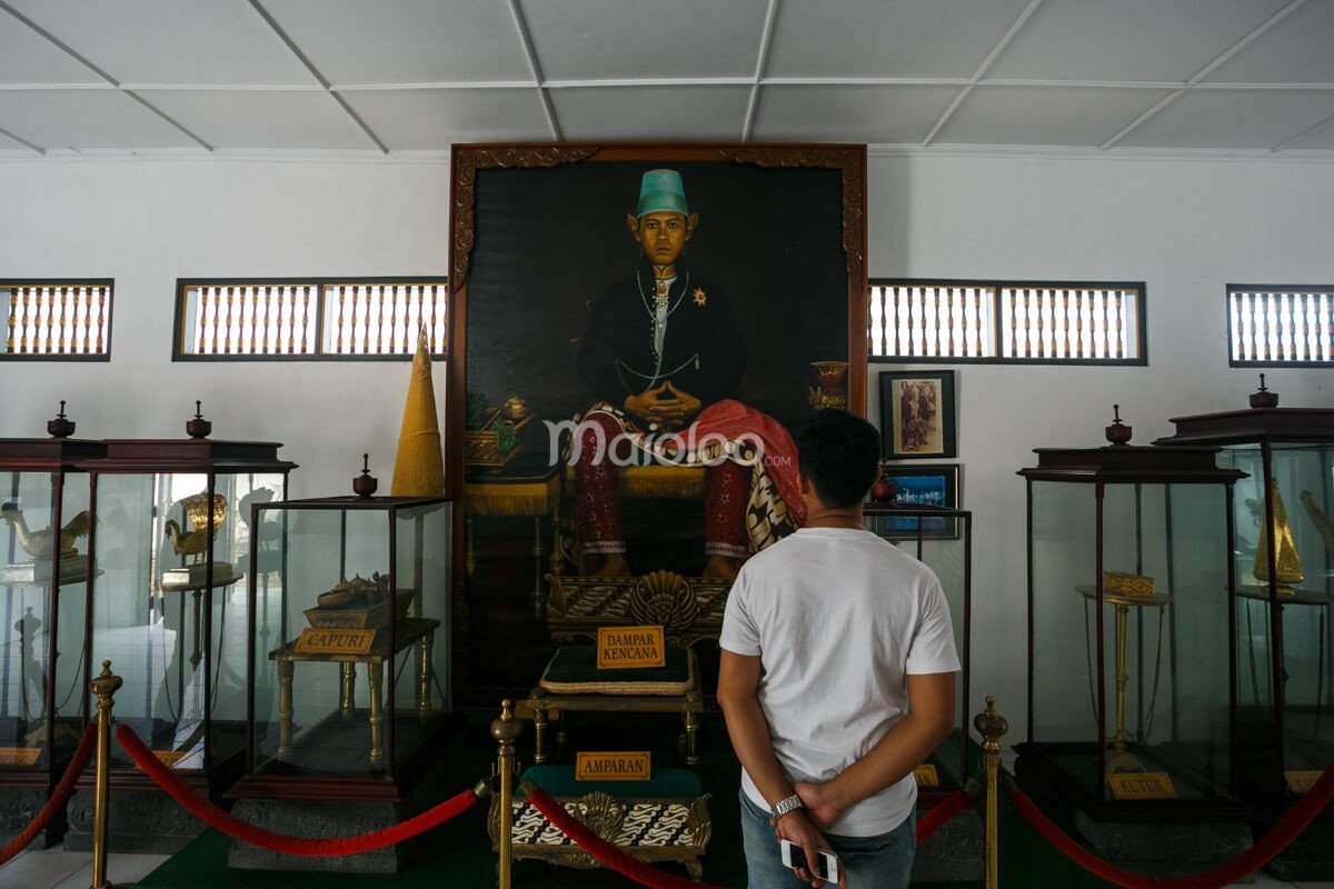 A visitor admiring a large portrait of Sultan Hamengkubuwono displayed in Yogyakarta Palace, surrounded by historical artifacts in glass cases.