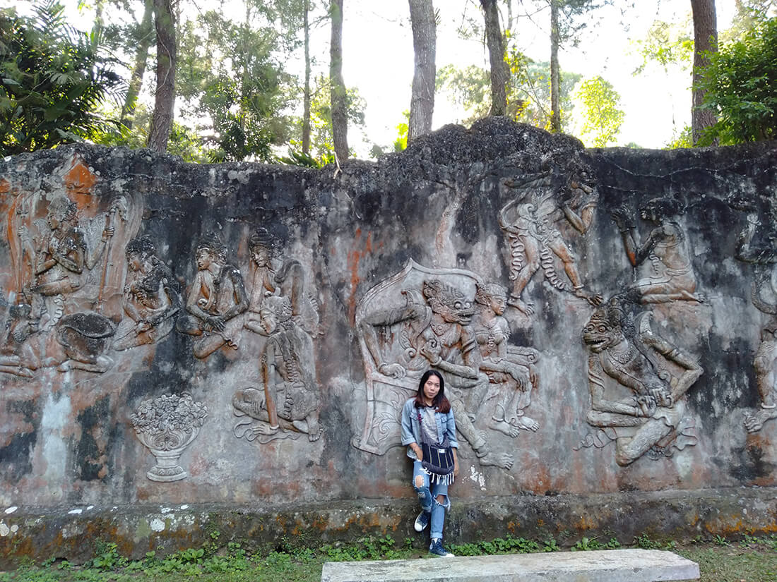 A person stands in front of a wall adorned with large and intricate reliefs depicting scenes from the ancient epic Ramayana, with surrounding trees adding to the setting.