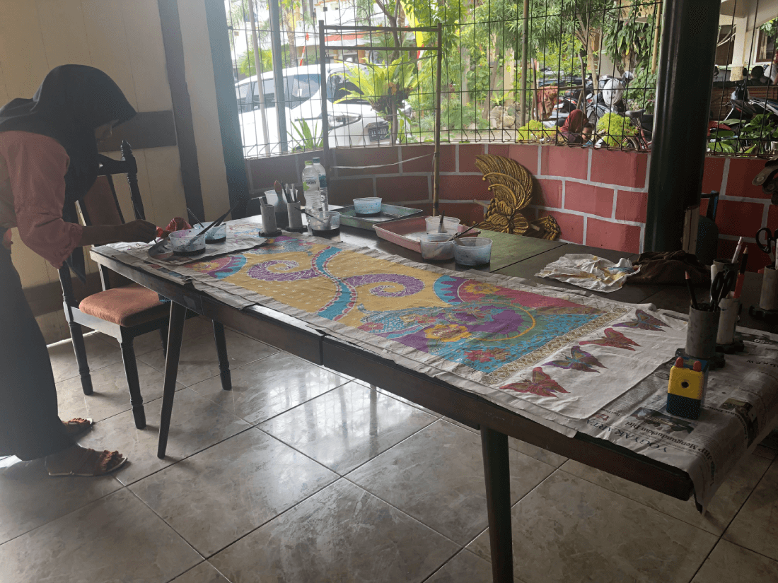 A woman working on a colorful batik fabric at a table in a workshop.