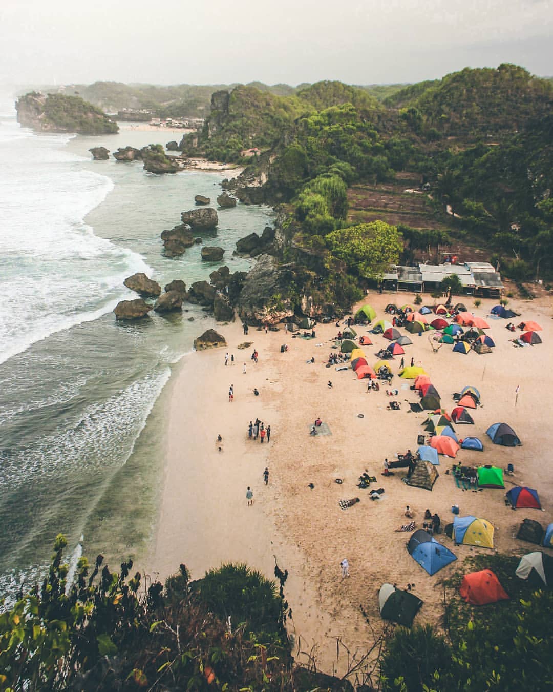 A bird's-eye view of Ngrumput Beach with multiple colorful tents set up on the sand, surrounded by rocky cliffs and lush green hills.