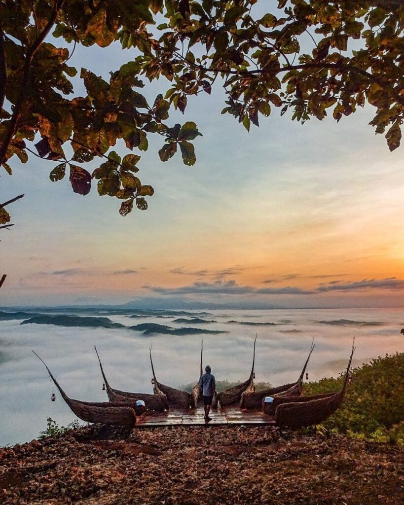 A person stands on a wooden platform with unique wicker structures, watching the sunrise over mist-covered hills at Geoforest Watu Payung Turunan.