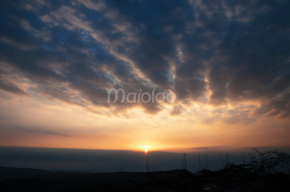 A stunning view of the sunset as seen from one side of Nglanggeran Mountain, with dramatic clouds and a golden sky.