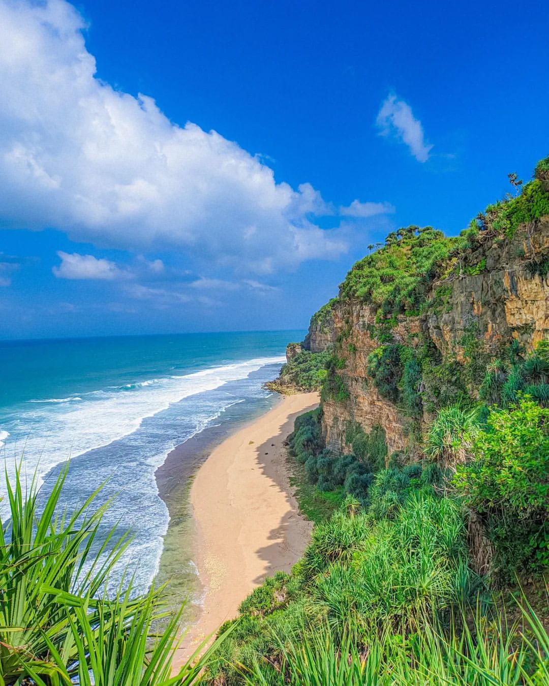 A breathtaking view of Watunene Beach with its sandy shoreline, turquoise waves, and towering karst cliffs under a blue sky.