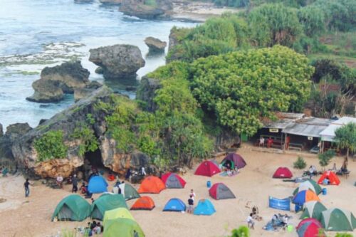 A beach with several colorful tents set up, surrounded by green hills and rocky cliffs, with the ocean in the background.