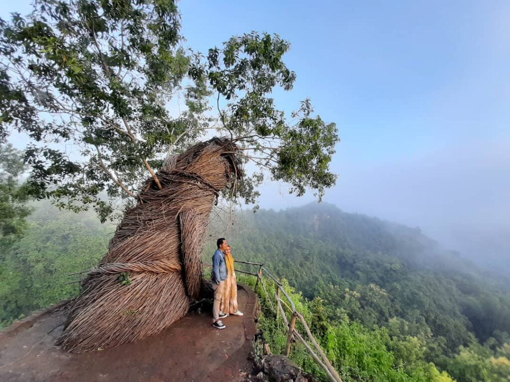 A couple stands in front of a large wicker installation wrapped around a tree on a cliff, with misty forest hills in the background at Geoforest Watu Payung Turunan.