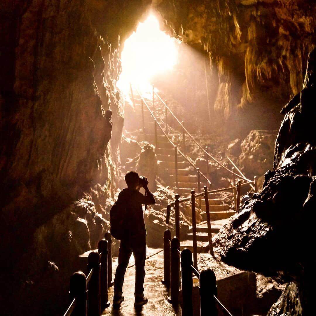 A person is taking a photo inside Kiskendo Cave, with sunlight streaming through the cave entrance and illuminating the rocky interior. Stairs lead up to the entrance.