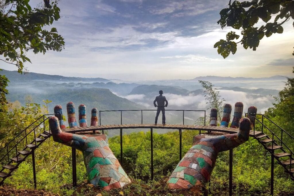 A person stands on a balcony supported by large colorful hand installations, overlooking misty valleys at Geoforest Watu Payung Turunan.