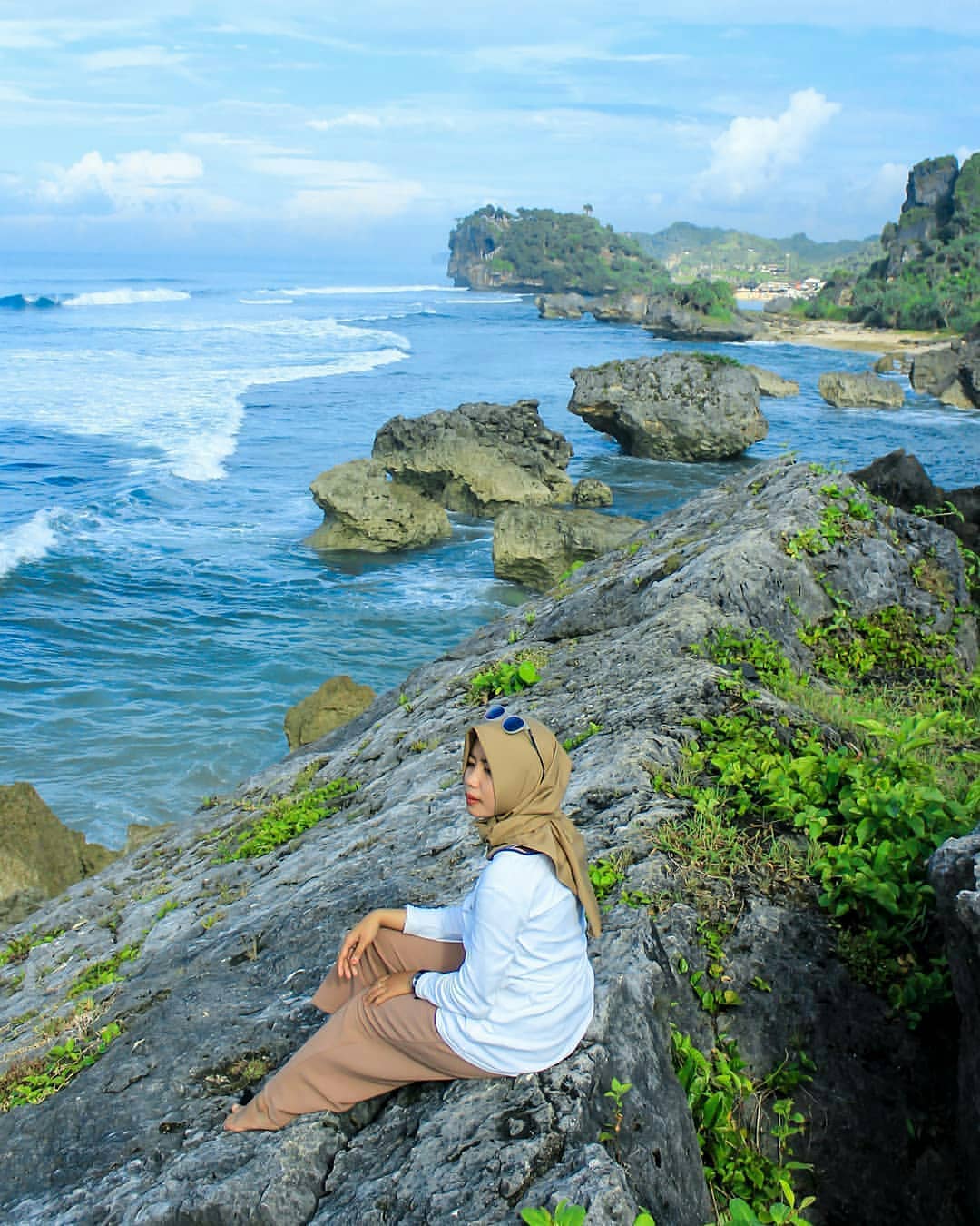 A woman sitting on rocky cliffs overlooking the ocean with waves crashing below and the coastline in the background.