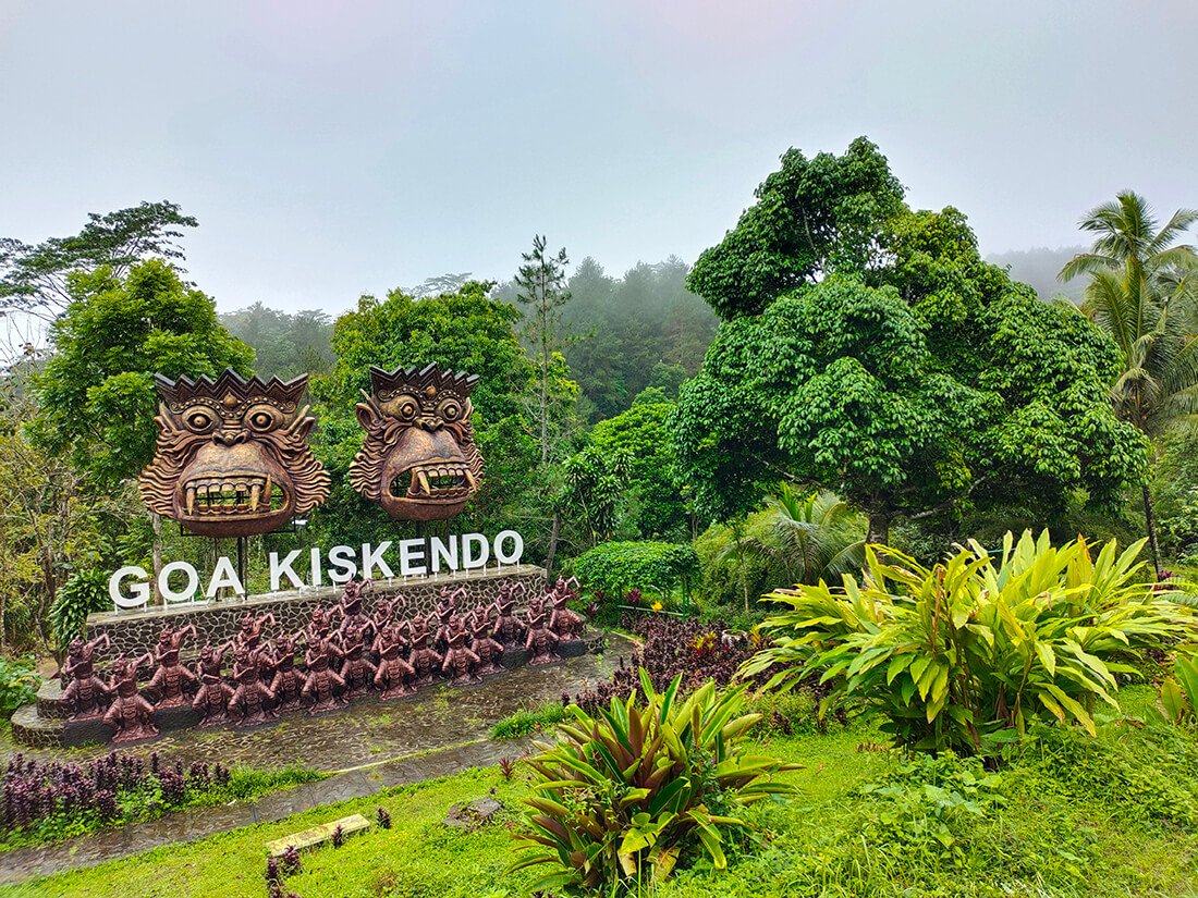 The entrance to Kiskendo Cave features two large decorative masks above a sign, surrounded by lush green vegetation and trees.