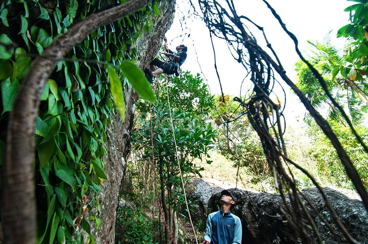 A climber scaling a rocky wall while another person oversees from the ground, surrounded by lush greenery on Nglanggeran Mountain.