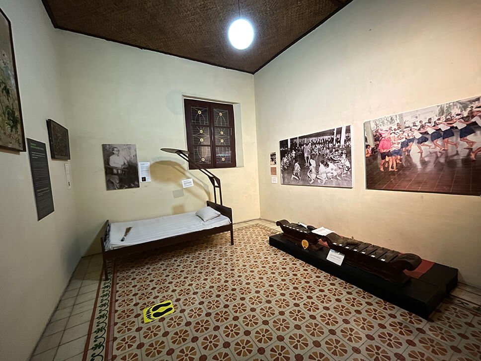 A room in Dewantara Kirti Griya Museum with a vintage bed, photographs, and a traditional musical instrument.