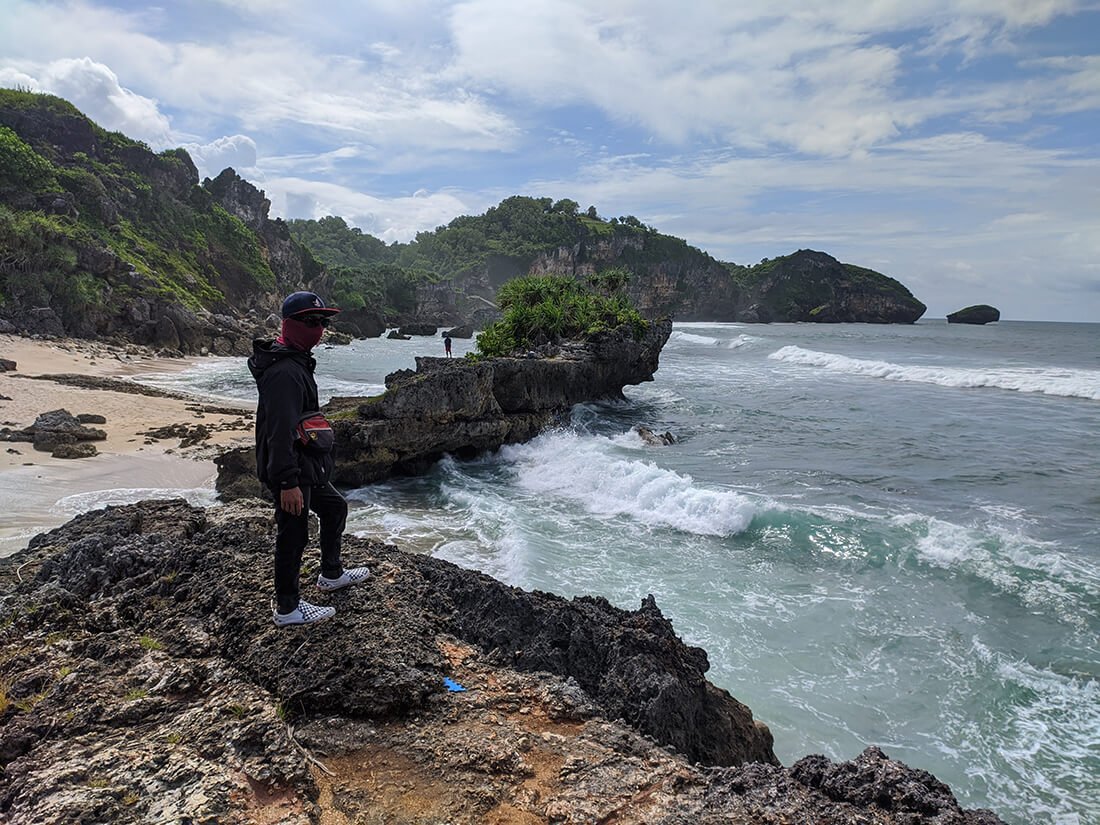 A person standing on a rocky shore overlooking the waves and cliffs at Watu Bolong Beach.