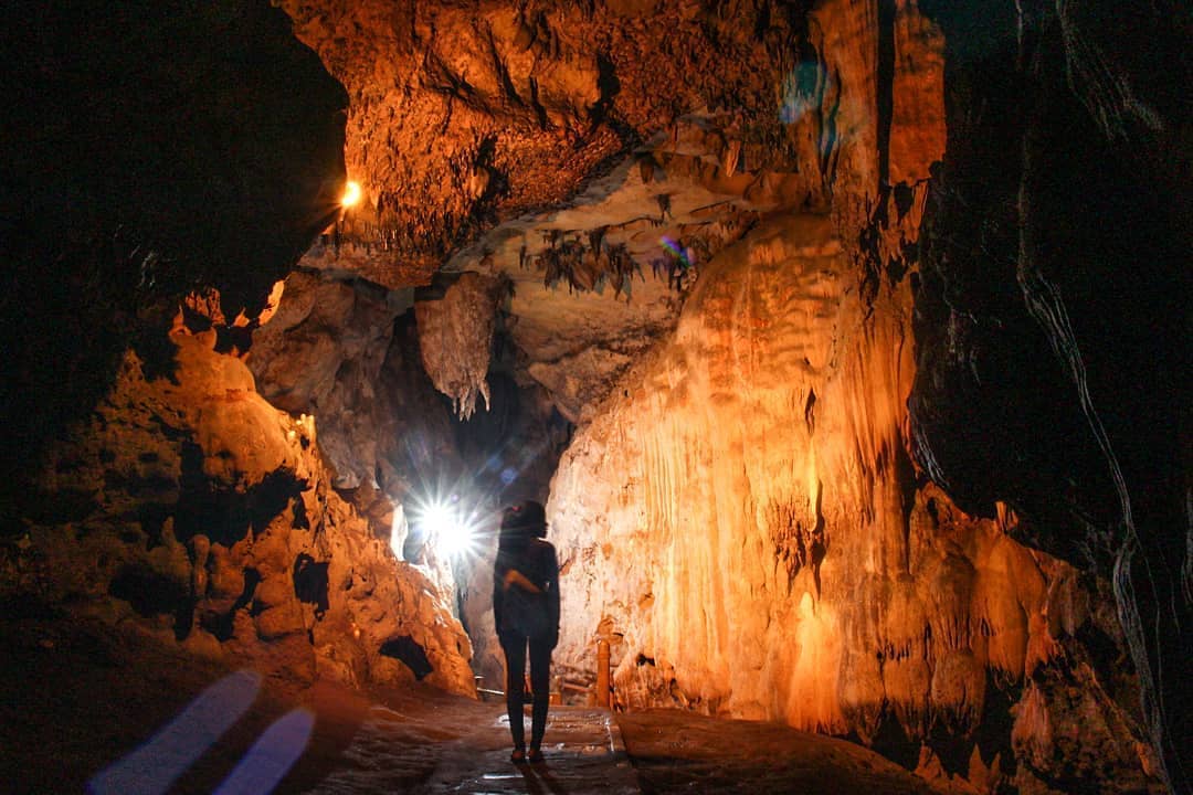 A person explores Kiskendo Cave, lit by flashlight, highlighting the intricate stalactite and stalagmite formations on the walls and ceiling of the cave.