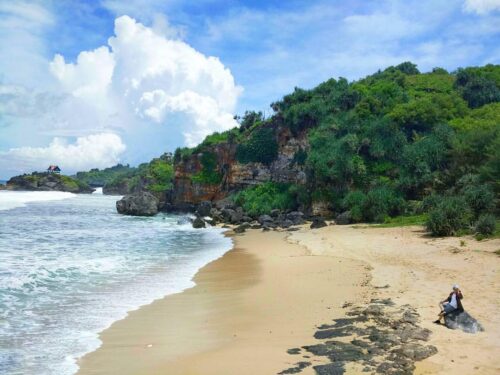 A serene beach with golden sand, rocky cliffs, and lush green vegetation under a clear blue sky.