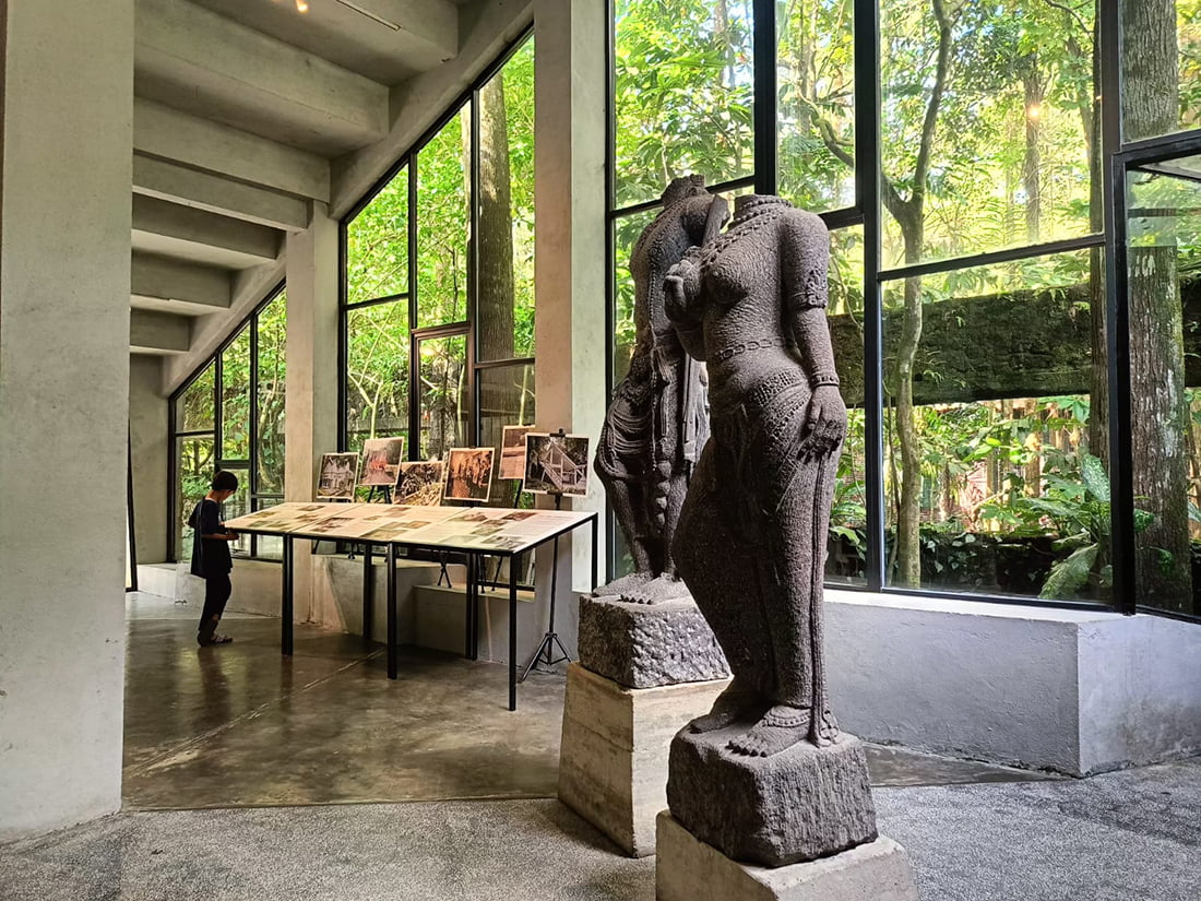 Stone statues and a person viewing historical photographs inside a gallery with large windows at Ullen Sentalu Museum.