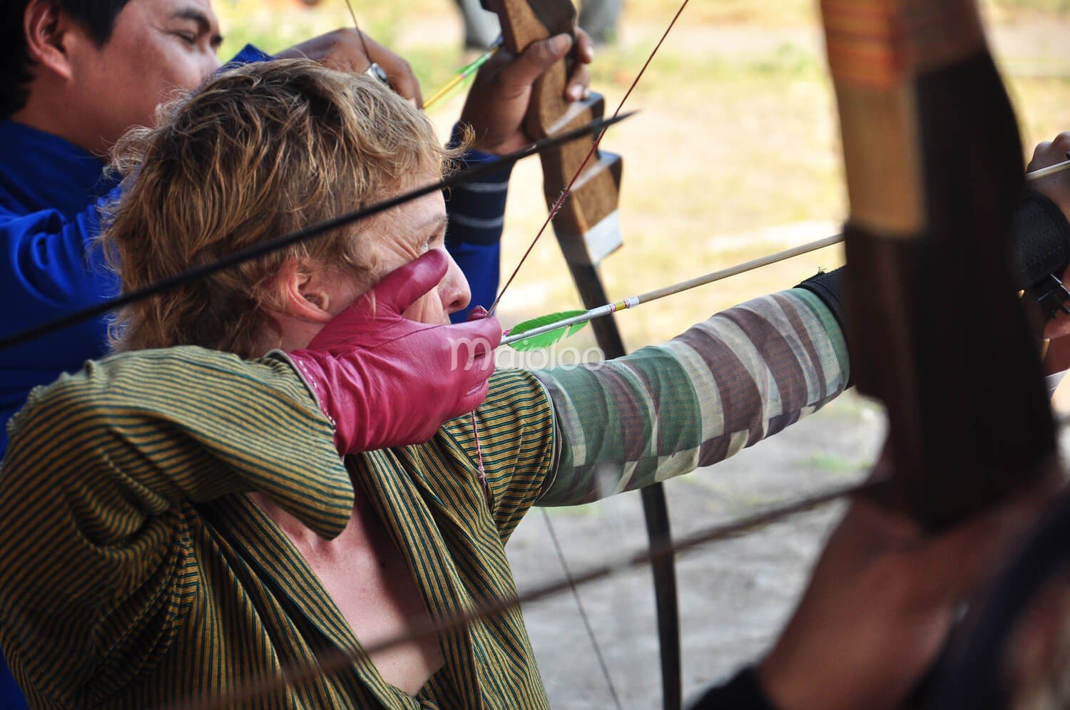 A person practicing traditional archery with a bow and arrow, wearing a striped outfit and pink glove.