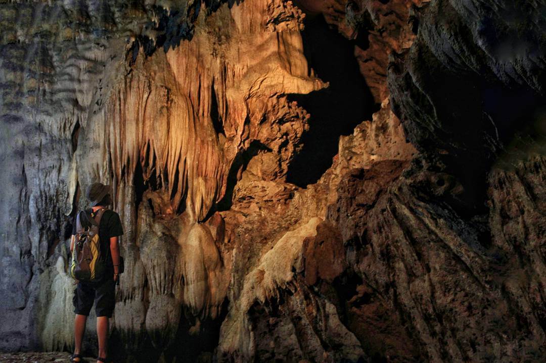 A person wearing a backpack and hat gazes at large stalactite formations on the wall inside Kiskendo Cave.