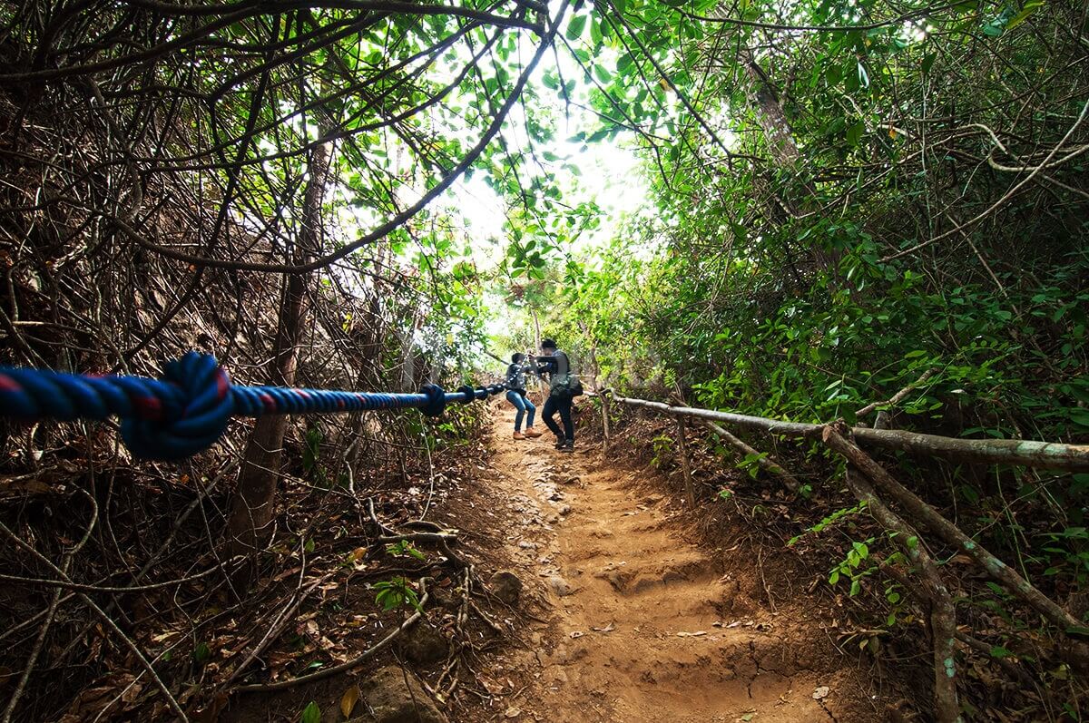 Two hikers holding onto a blue rope as they navigate a steep and forested trail on Nglanggeran Mountain.