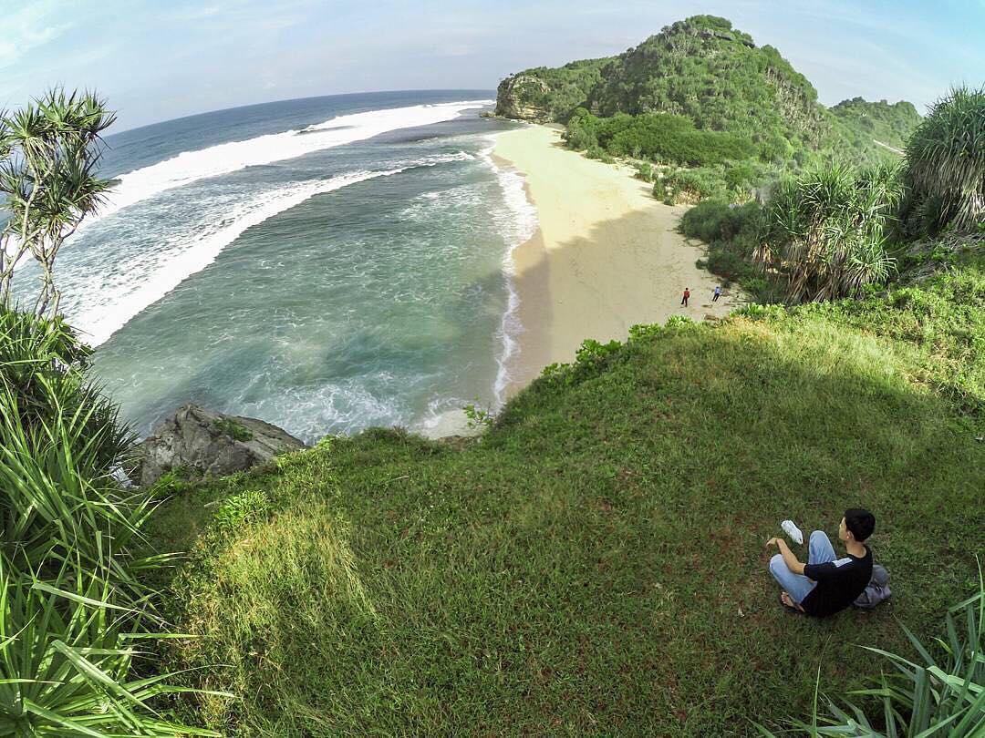 A person sitting on a grassy hill overlooking Sanglen Beach with its white sand and clear blue water.