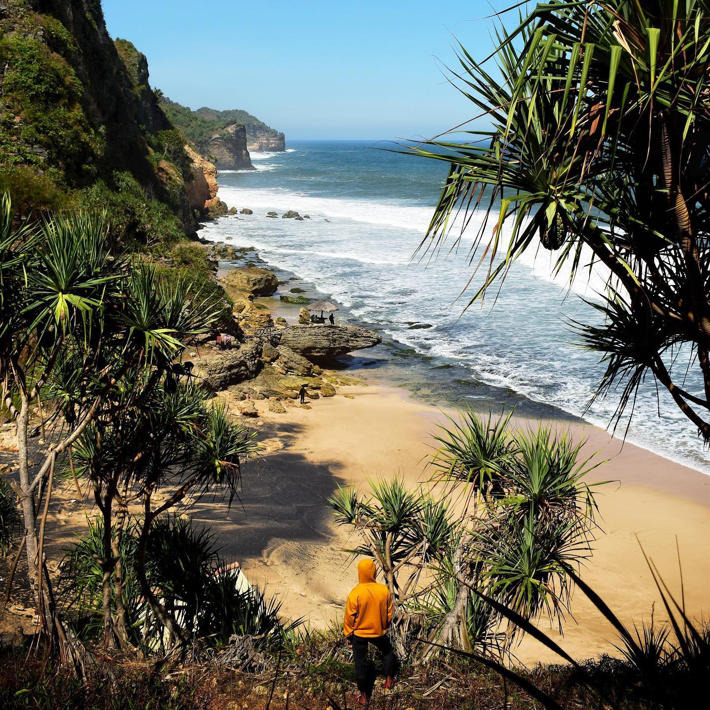 A person in an orange hoodie stands on a cliffside overlooking Seruni Beach with waves crashing onto the sandy shore below.