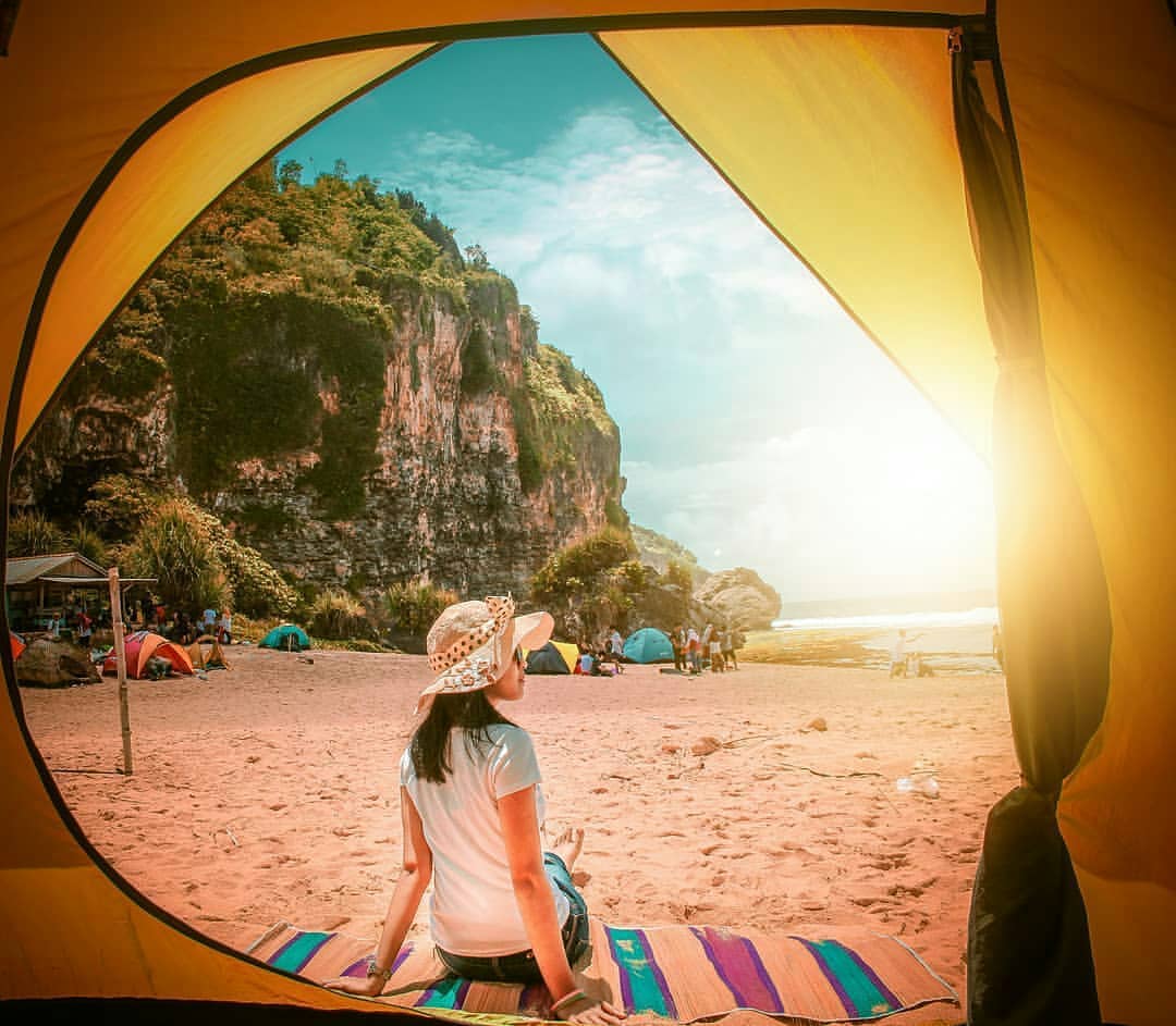 A woman sitting inside a tent looking out at the beach with cliffs, other tents, and people in the background during sunset.