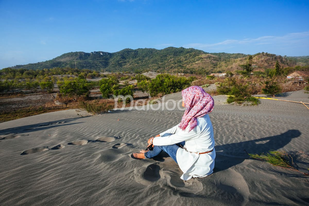 A person sits on the sand, enjoying the view of hills and greenery at the dunes.