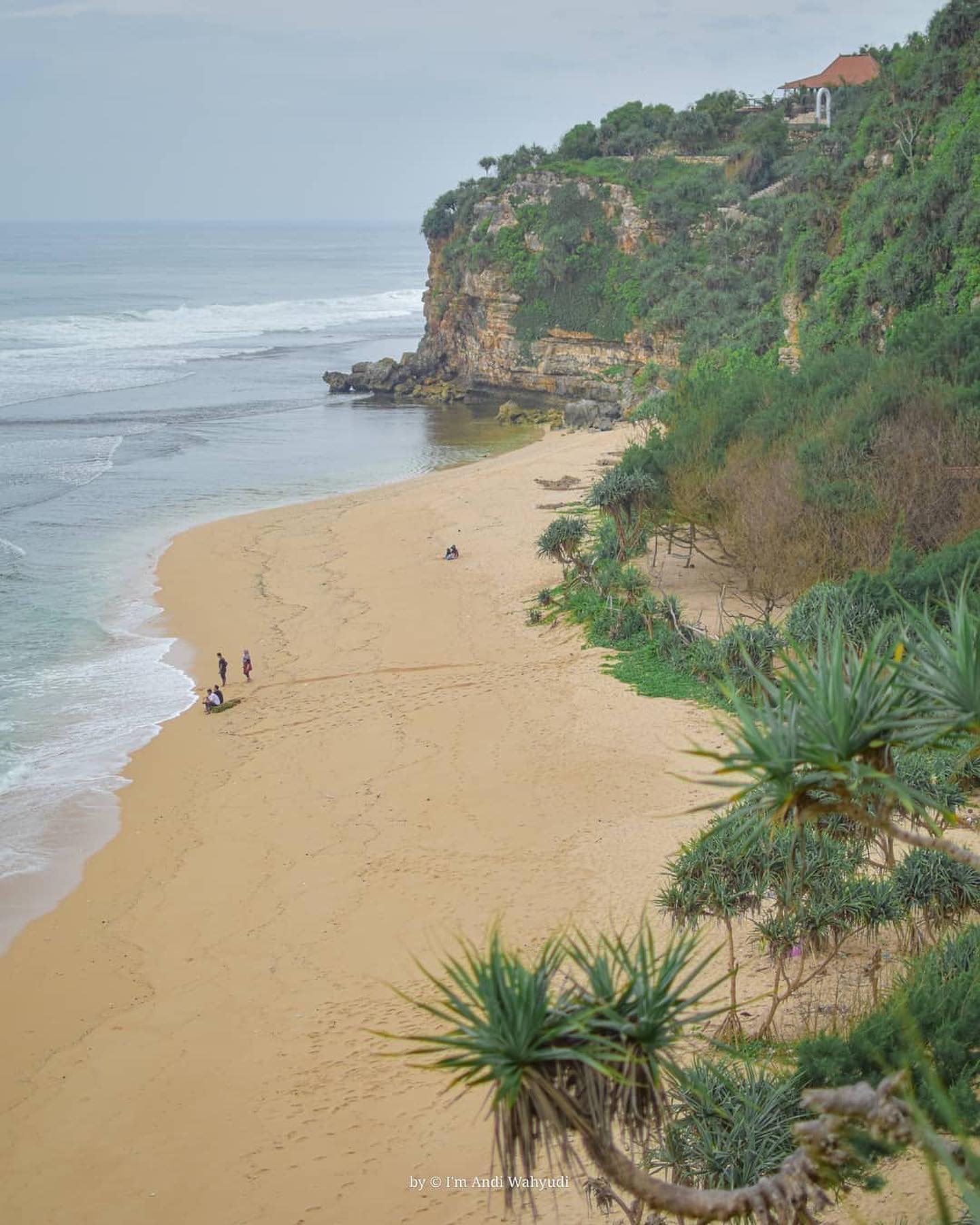 A calm day at Sanglen Beach with a few people walking along the sandy shore near a cliff covered in greenery.