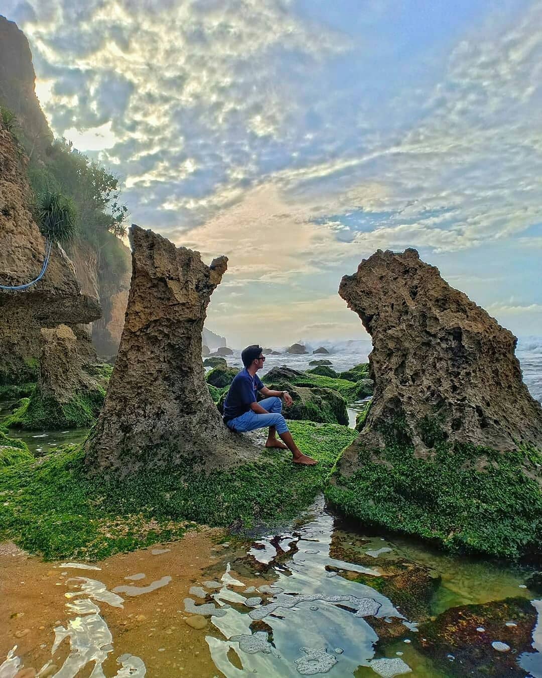 A person sits between two large rock formations covered with green seaweed at Seruni Beach, gazing at the ocean.