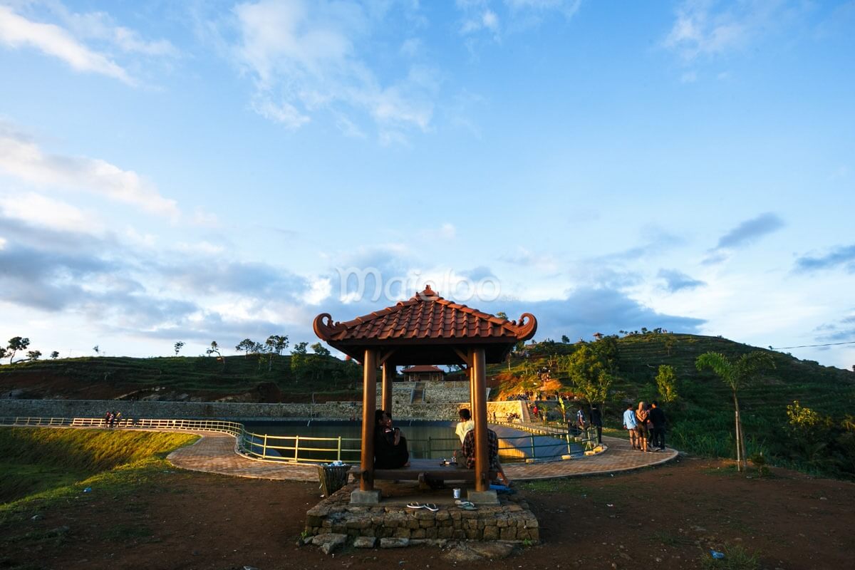 A small wooden gazebo with a tiled roof by a reservoir, with people sitting inside and enjoying the view.